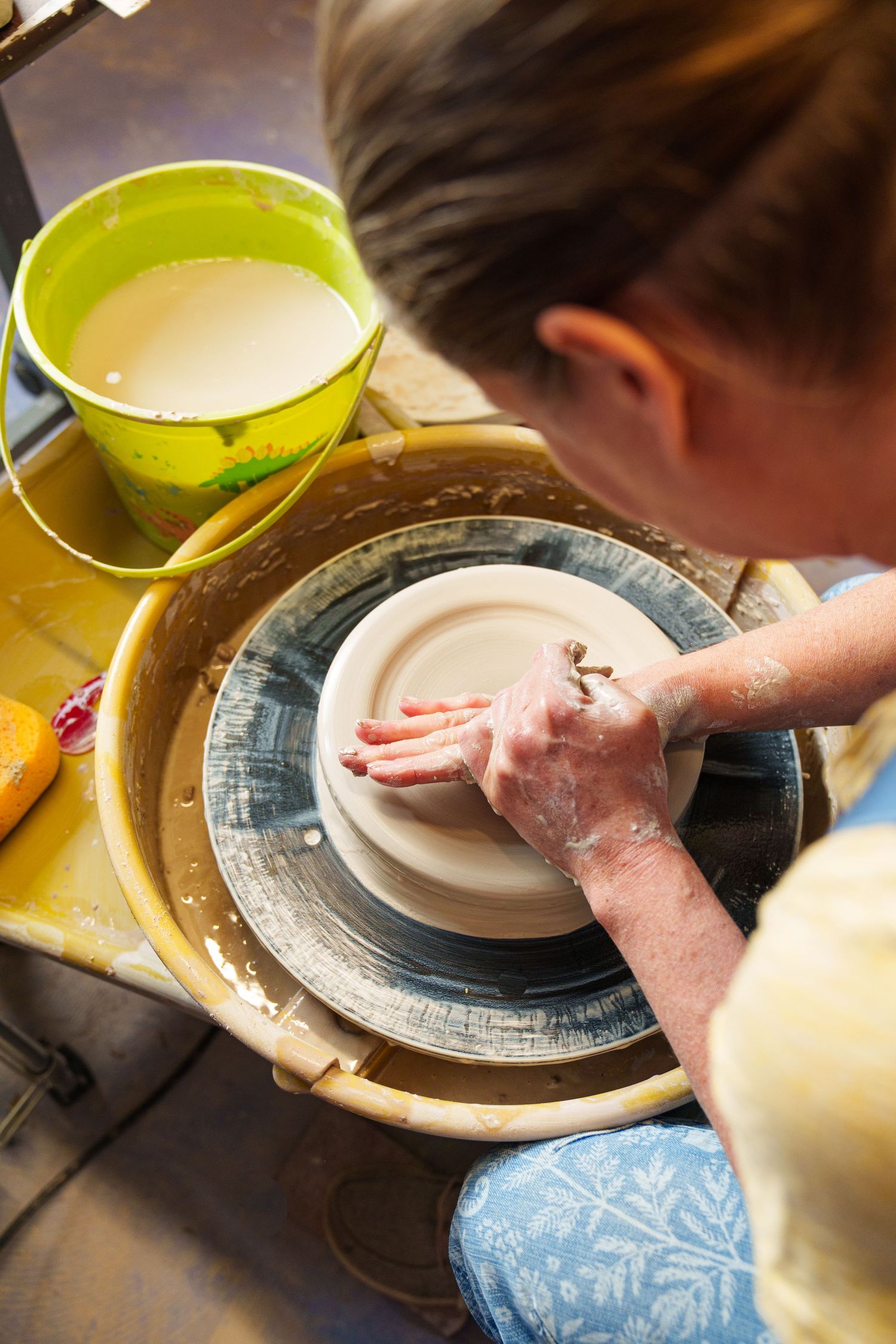 Close-up of hands working clay on a pottery wheel with yellow tools and blue surface.