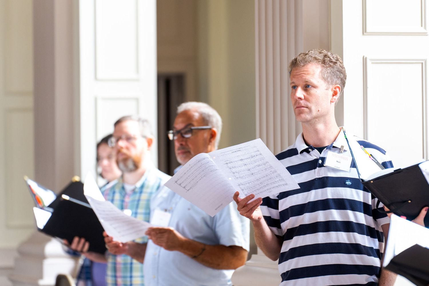 A group of people in striped shirts stand together in a bright hallway looking at papers and documents.