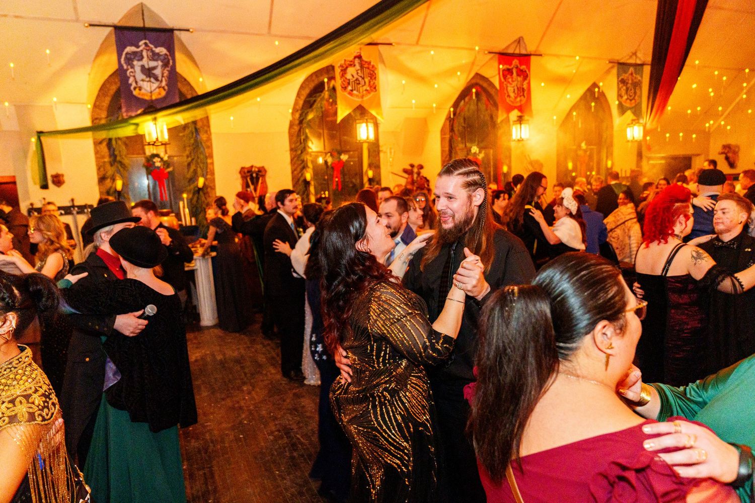 Couples dancing together at an elegant evening event in a warmly lit ballroom with arched windows.