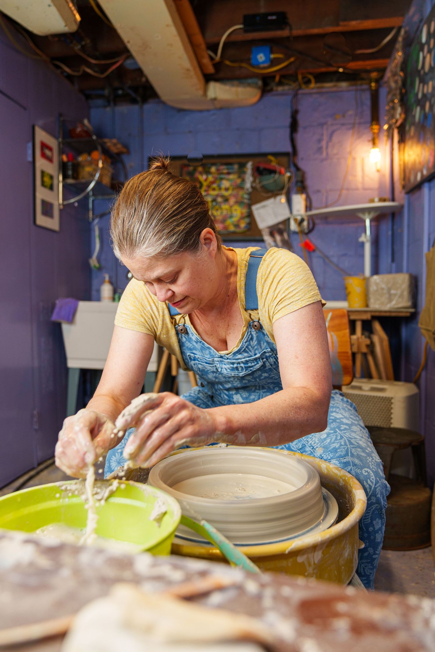 A potter works at a pottery wheel shaping clay bowls in a dimly lit studio with blue walls and hanging lights.
