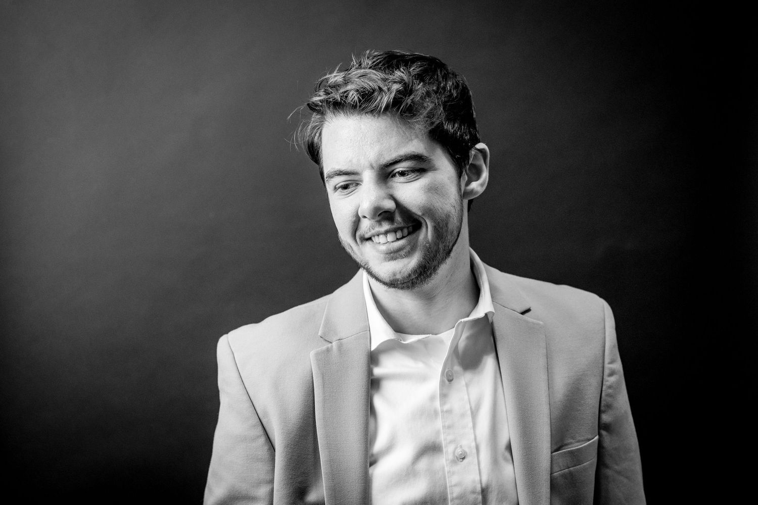 A black and white professional portrait shows someone in a light colored suit jacket smiling against a dark background.