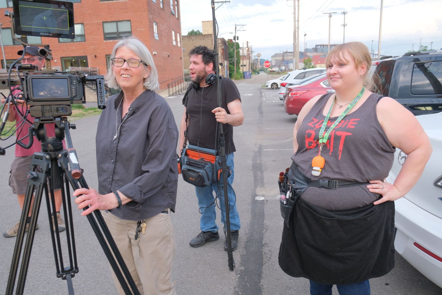 Three people stand outdoors near a video camera on a tripod, filming on a city street.