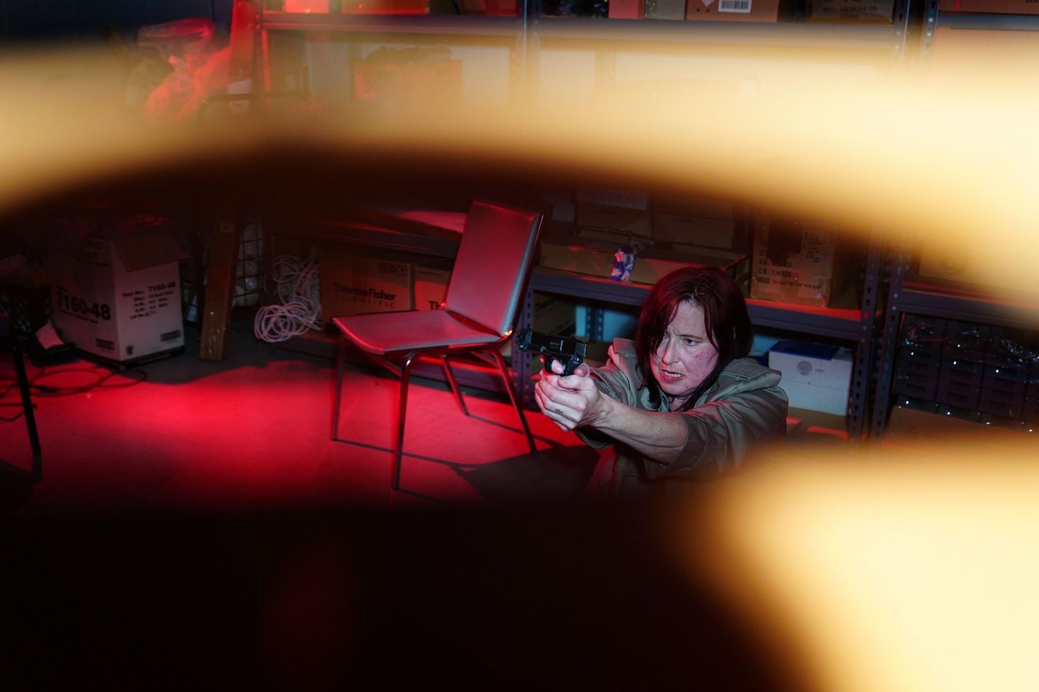 Woman aiming a handgun in a dimly lit room with red lighting and office chairs in the background.