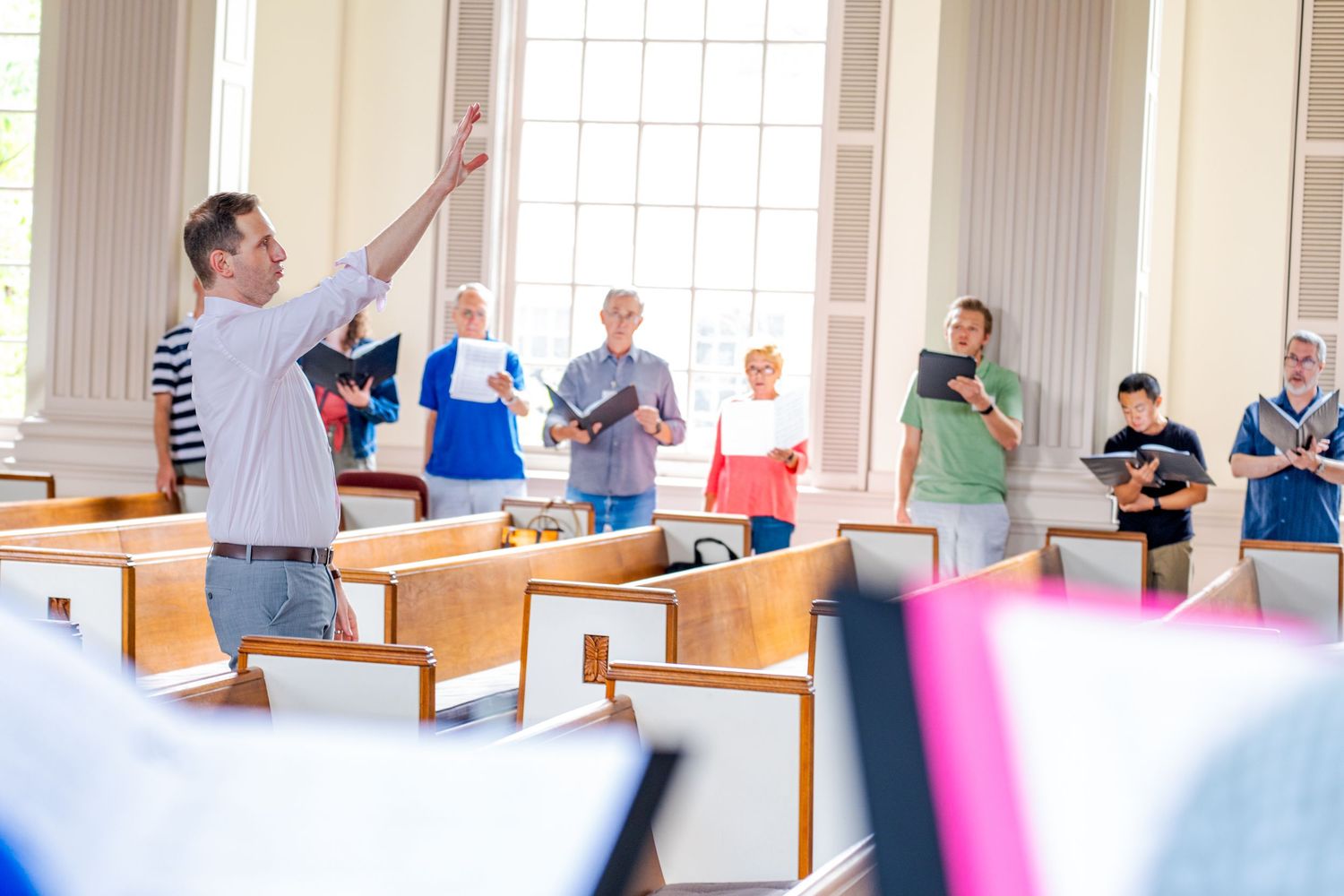 A choir director leads a group of singers rehearsing in a bright church sanctuary with wooden pews.