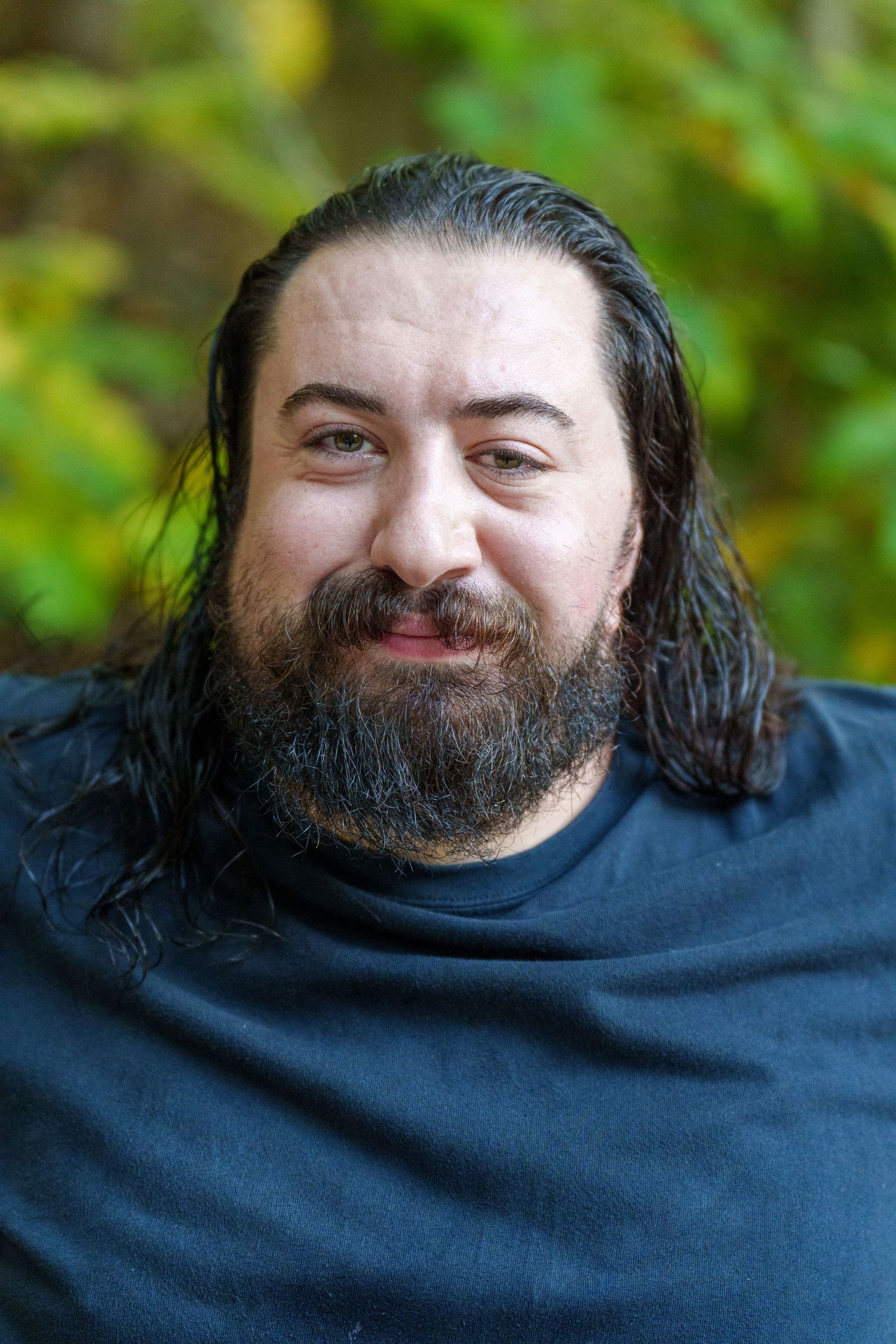 Smiling bearded man with long dark hair wearing a black shirt outdoors with green foliage background.
