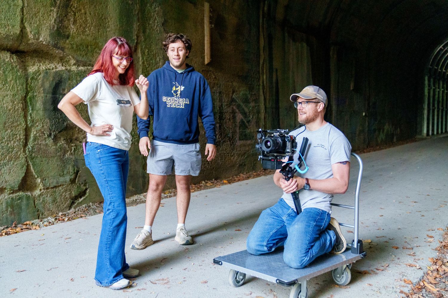 Filmmaker on dolly films two young adults near a tunnel outdoors during a video production shoot.