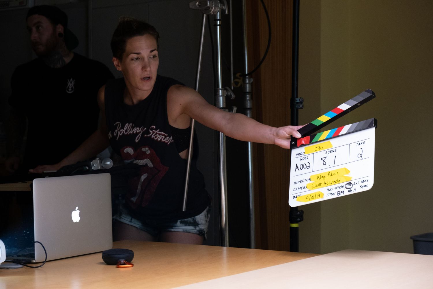 Woman in Rolling Stones shirt holds a film clapperboard on a movie set with a laptop nearby.