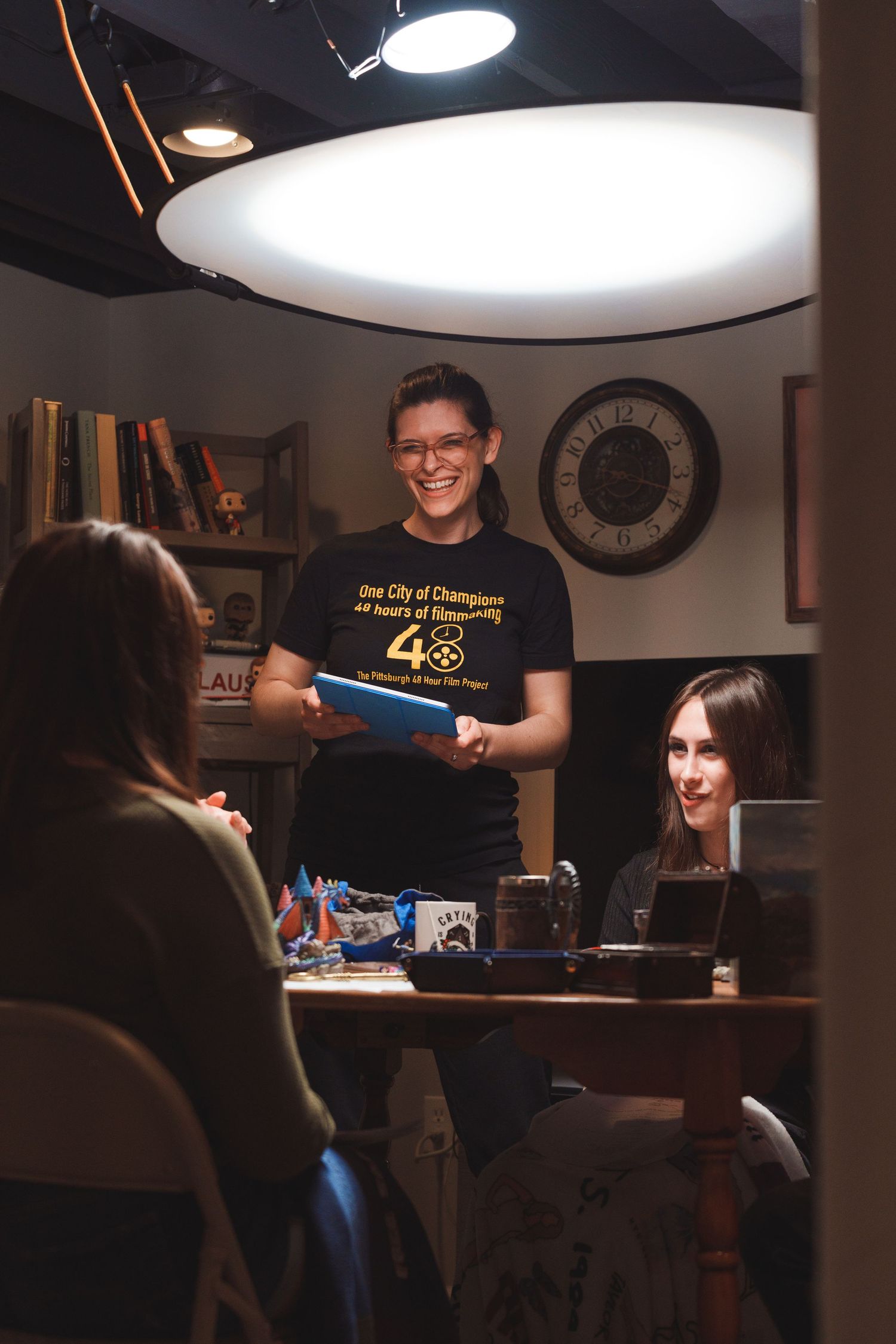 Woman in a black Champions t-shirt smiling while holding a folder under bright studio lighting with colleagues.