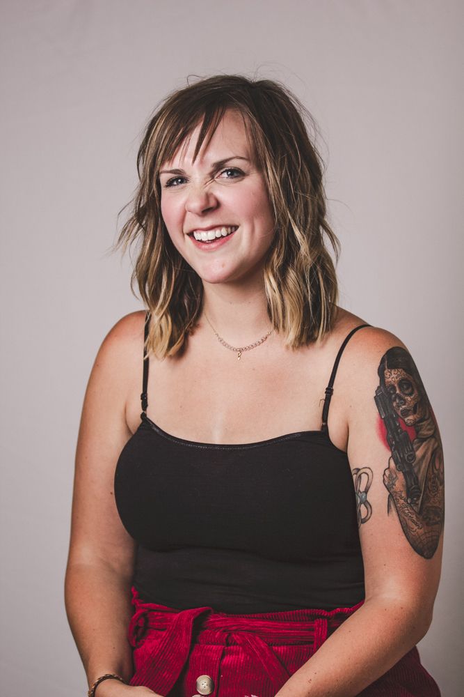 Portrait of someone in a black tank top and red skirt with visible arm tattoos smiling against a gray background.