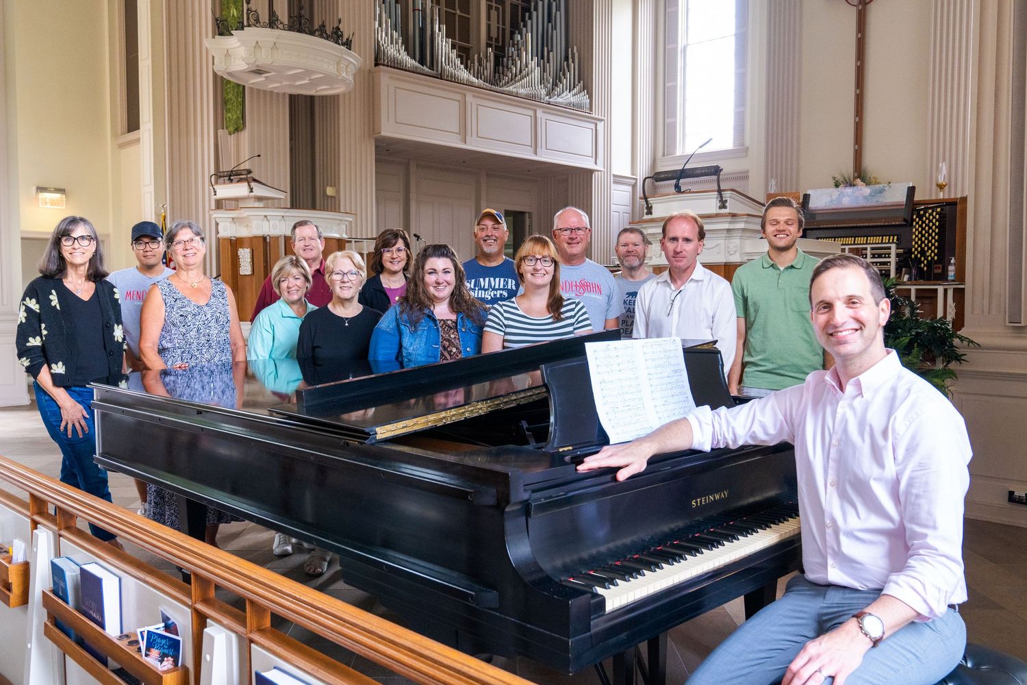 Group of people sitting in church pews next to a piano during a musical performance or rehearsal.