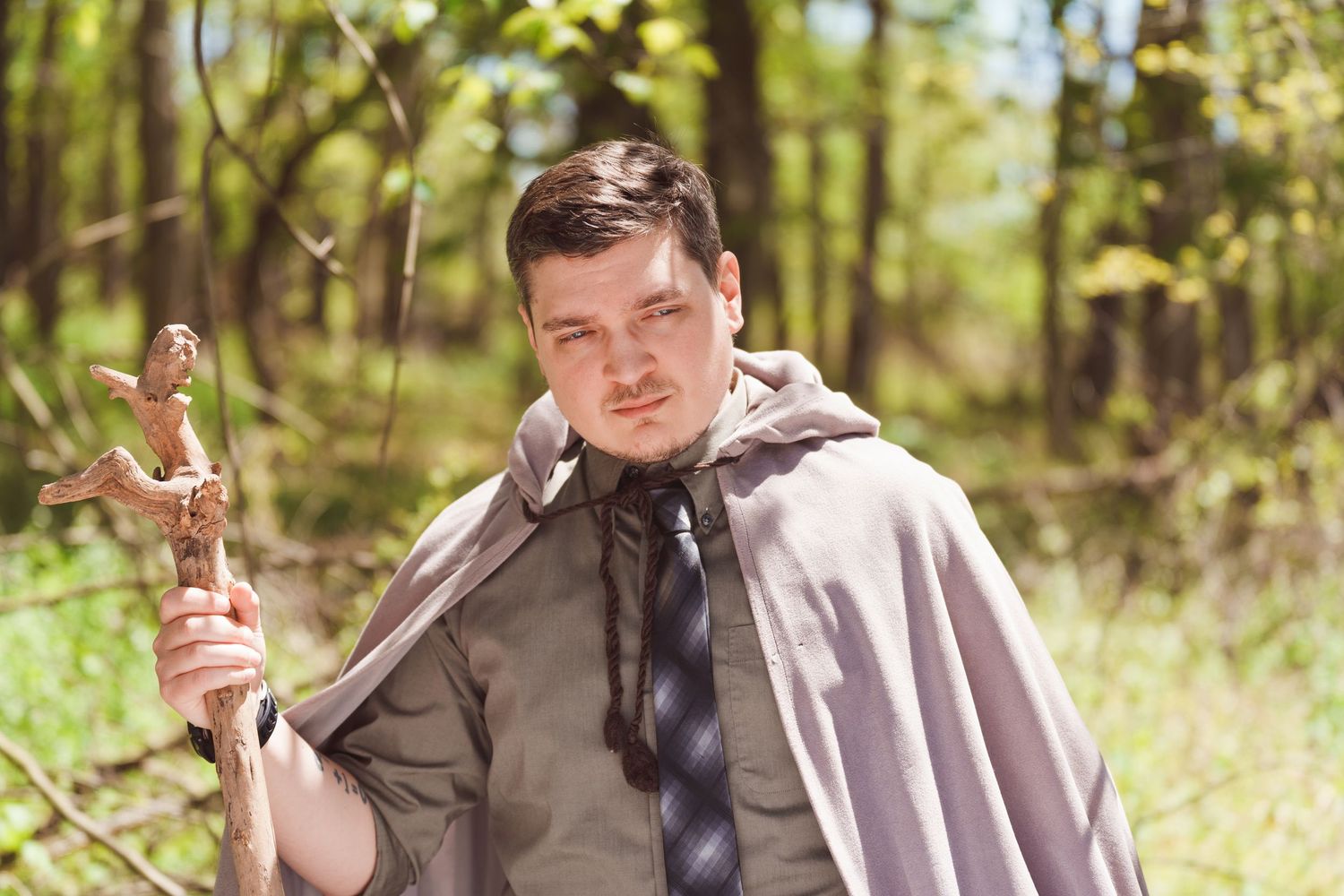 Man in gray cape holding wooden cross staff in sunlit forest, cosplay or fantasy roleplay outdoor portrait.