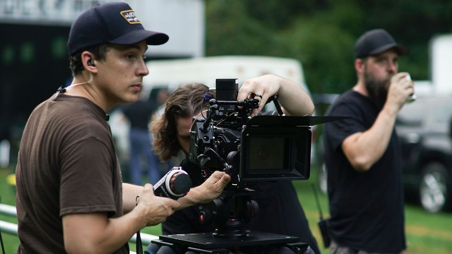 Film crew operating a camera on an outdoor set with green trees in the background.