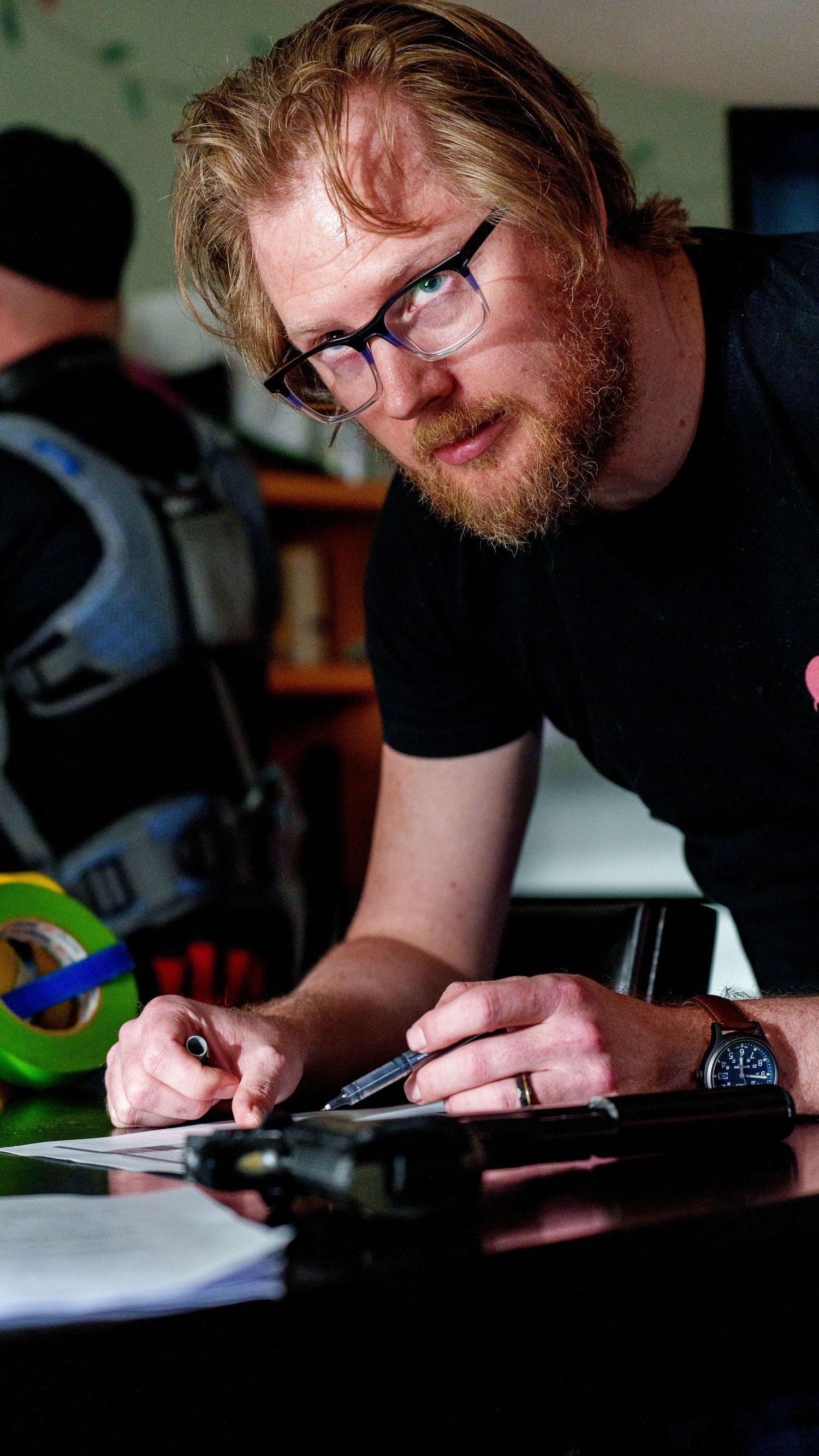 Person with glasses and beard working at desk with writing materials and office supplies in dark lit room.