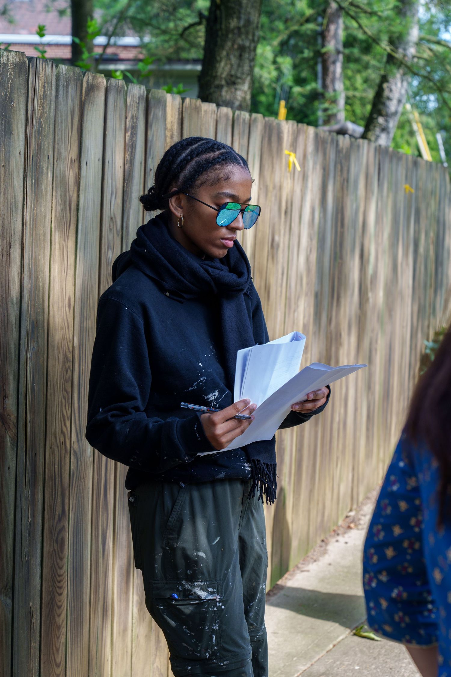 Woman in black hoodie and green sunglasses reading papers outdoors near a wooden fence.