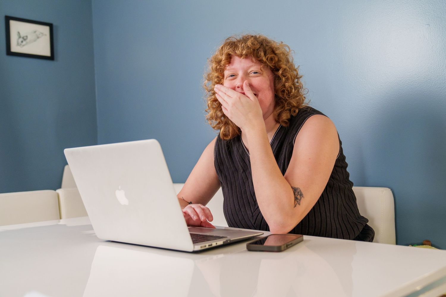 Curly red-haired woman covering mouth with hand while working on Apple MacBook laptop at white desk.