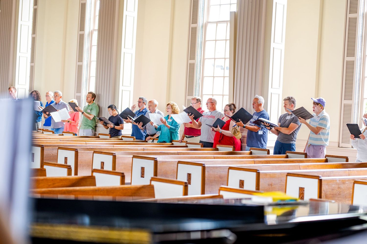 Large choir group rehearsing with sheet music in a bright church with wooden pews.