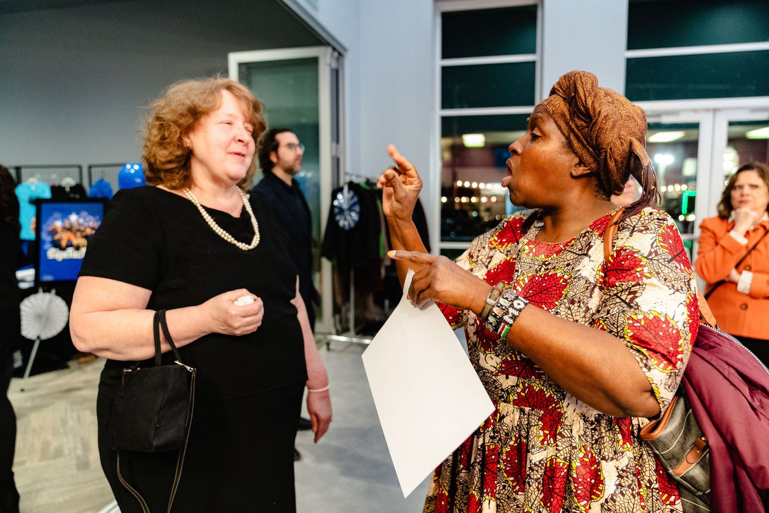 Two women share an animated conversation while laughing together at an indoor gathering.
