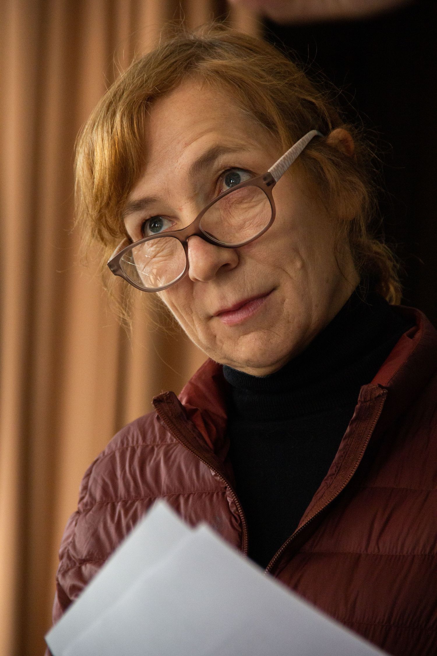 Middle-aged woman with red hair and glasses holding papers, wearing a burgundy vest over black turtleneck.