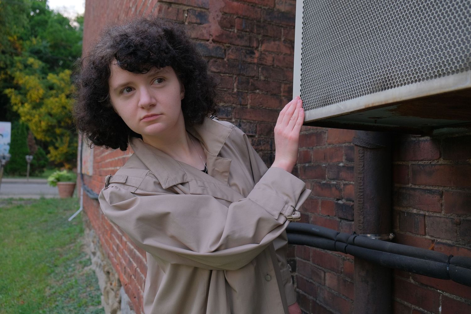 Young woman with curly hair in beige trench coat leaning against brick wall outdoors.