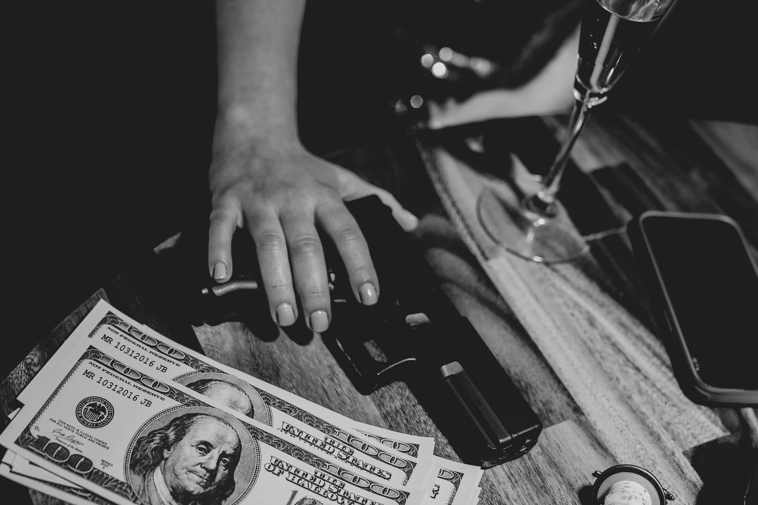 Black and white close-up of hand reaching for cash dollars on wooden surface near smartphone.