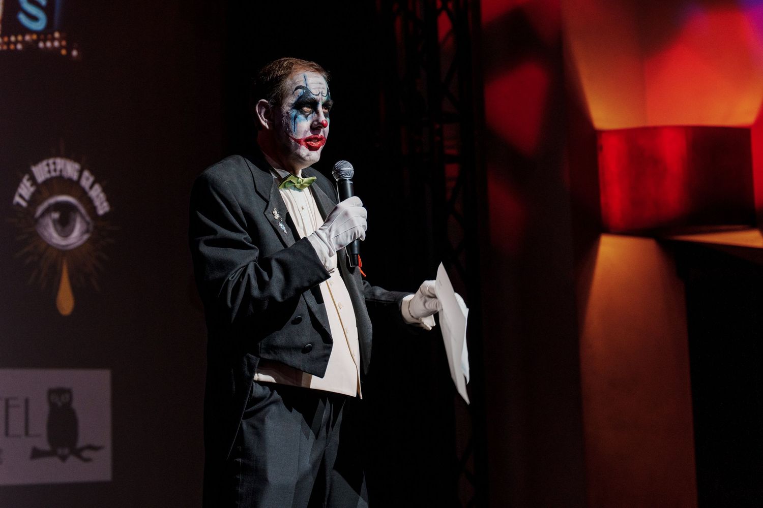 Performer in clown makeup speaks into microphone on dimly lit stage.