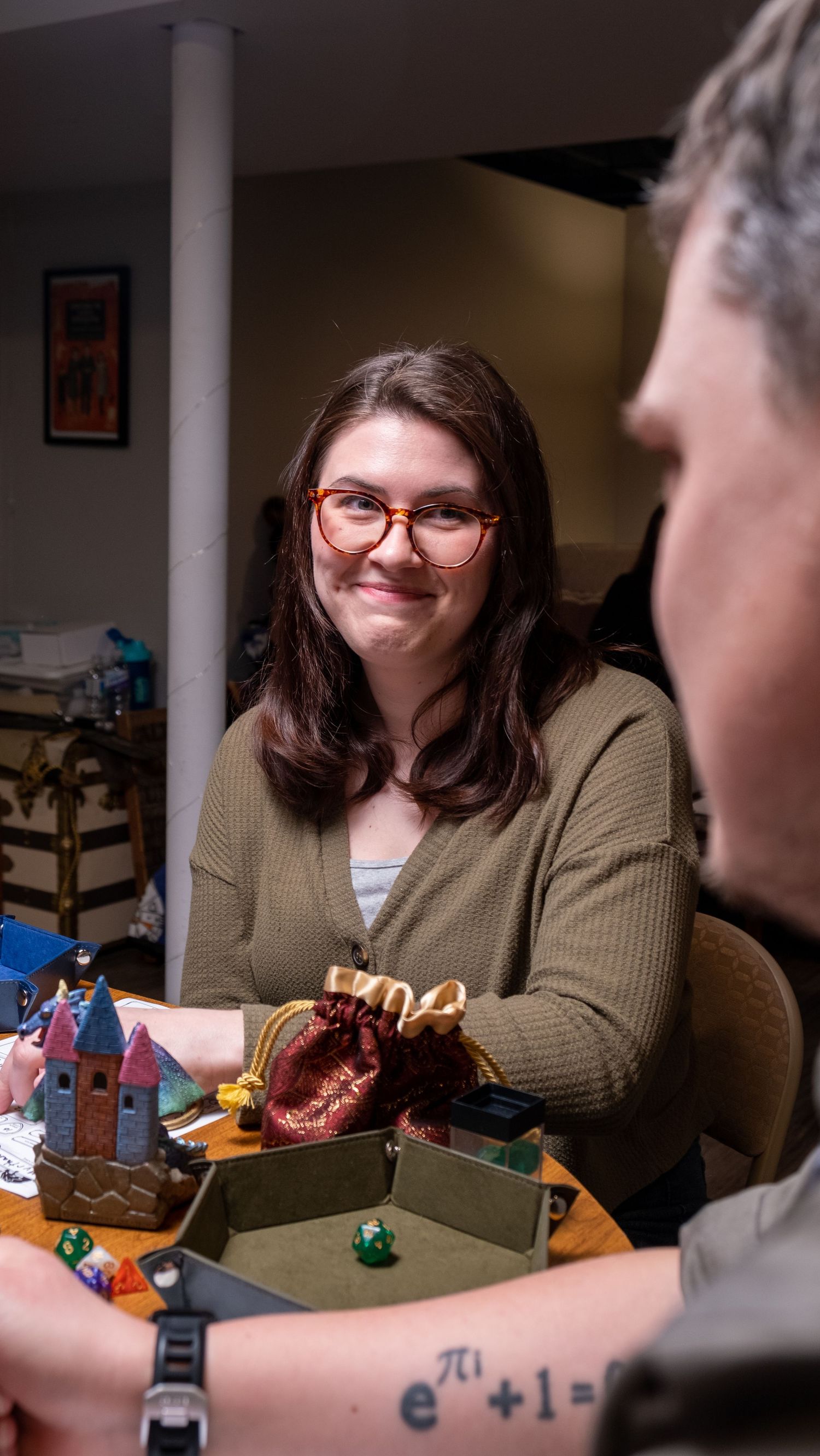 Young woman with glasses smiles at a tabletop RPG game session with dice and miniatures on the table.