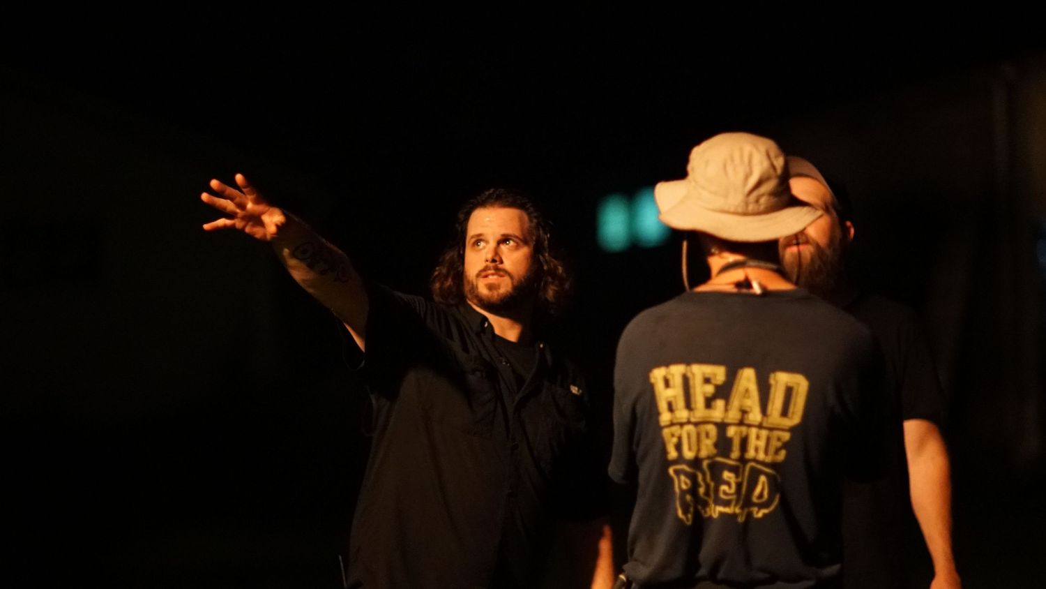 Two men in dark setting, one pointing and wearing black, other in HEAD FOR THE HILLS t-shirt and hat.