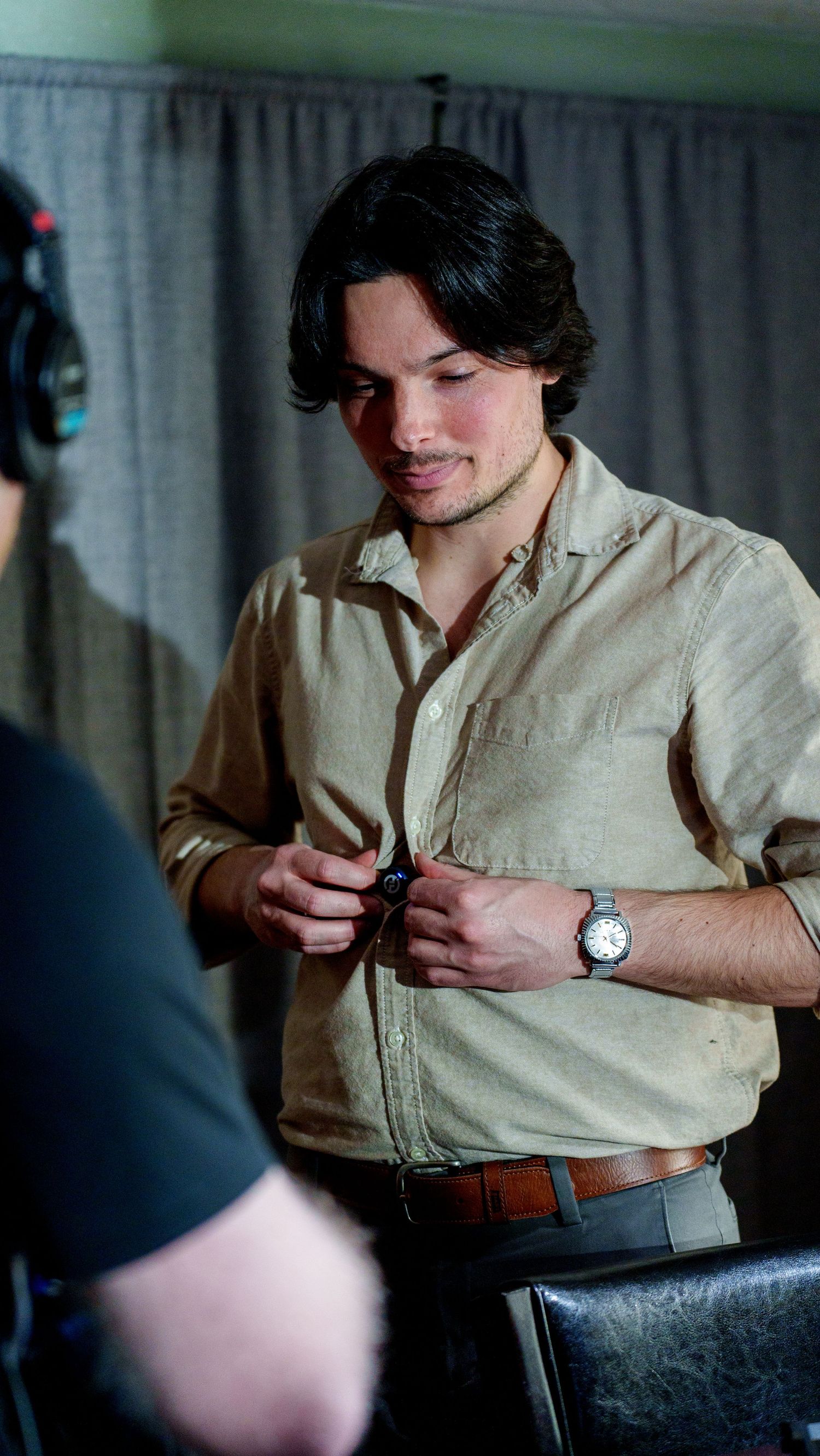 A young man with dark hair wearing a beige linen shirt and silver watch adjusts his clothing indoors.