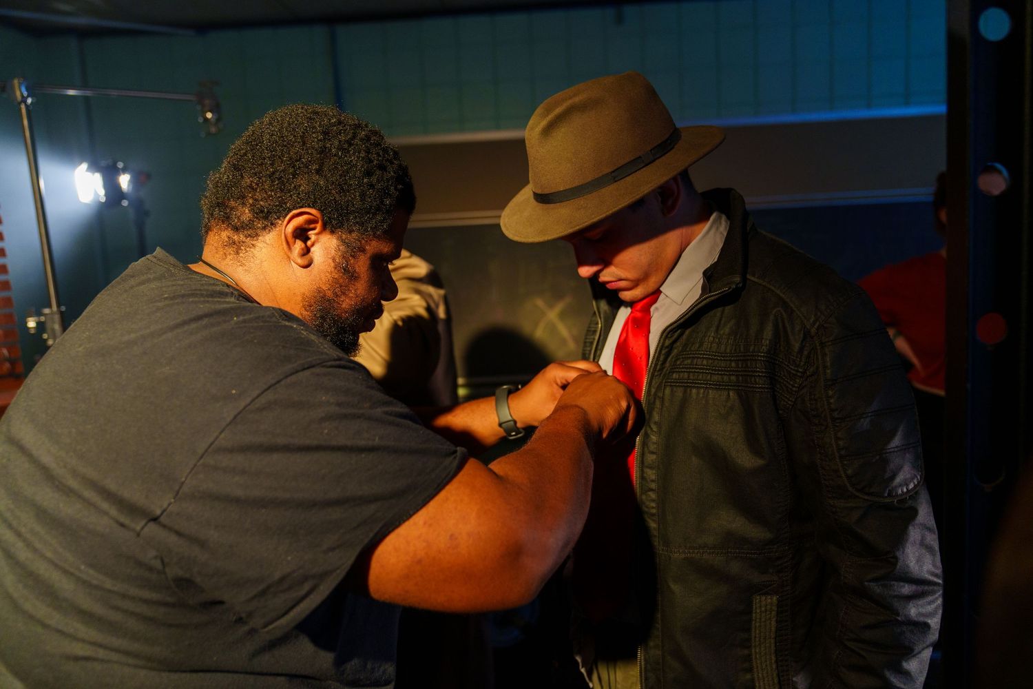 Two men interact closely indoors, one wearing a tan fedora, red tie, and olive jacket under dim warm lighting.