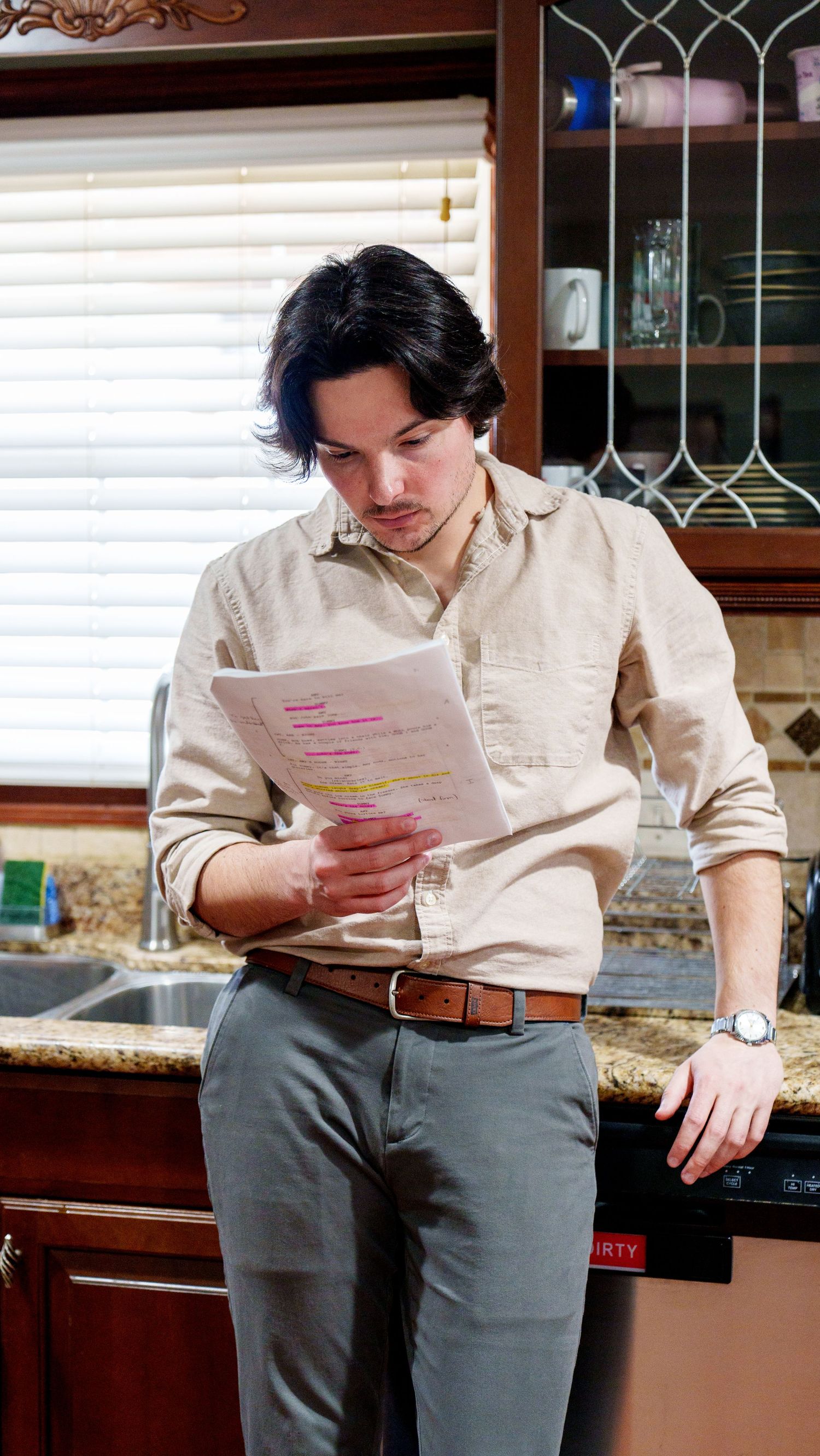 Person in beige shirt and gray pants reading documents while standing in a kitchen setting.