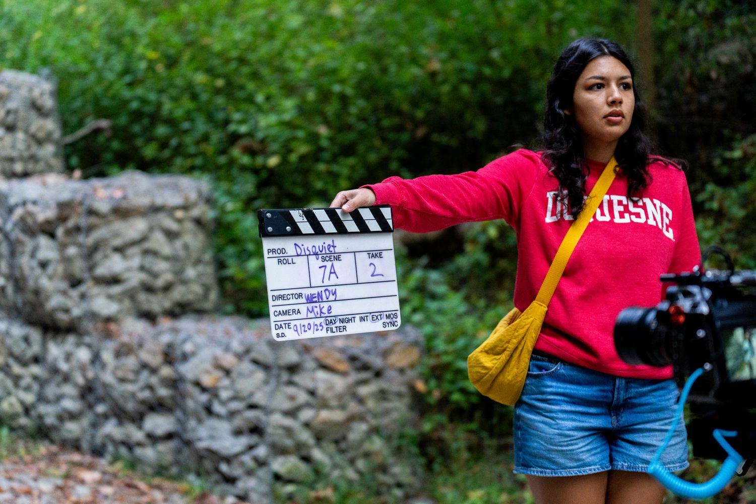 A film crew member in a red sweatshirt and denim shorts holds a movie clapperboard outdoors against a stone wall background.