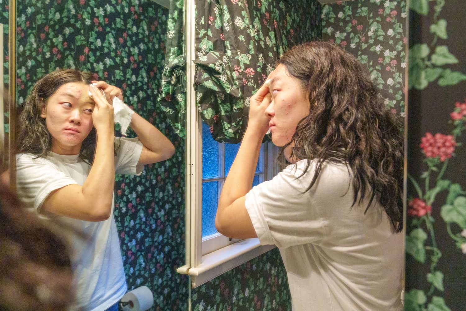 Woman examining her face in a bathroom mirror with floral wallpaper background.