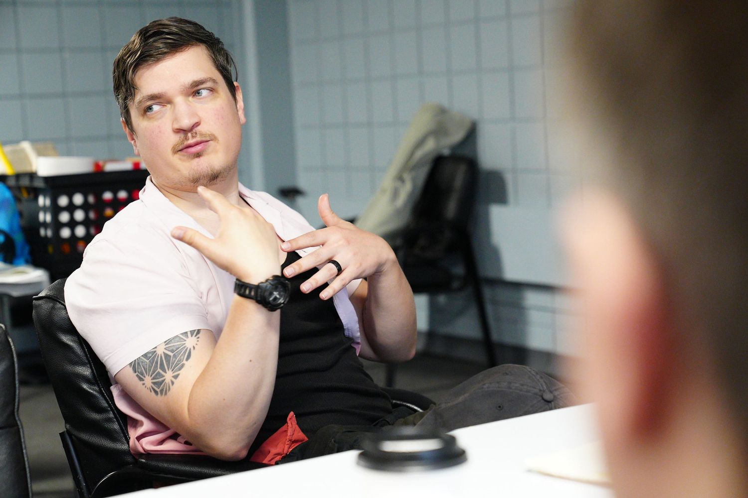 A tattooed man in a pink shirt gestures while speaking during a casual office meeting.