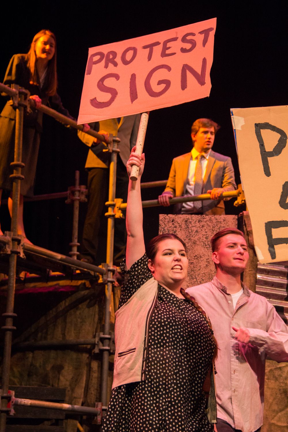 Actors hold pink protest signs during dramatic stage performance.