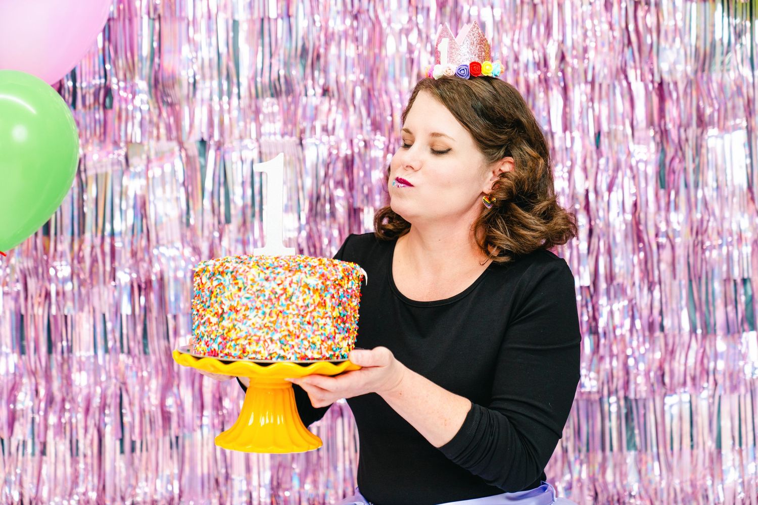 Woman in black top enjoying cake in front of sparkly pink backdrop at a festive celebration.