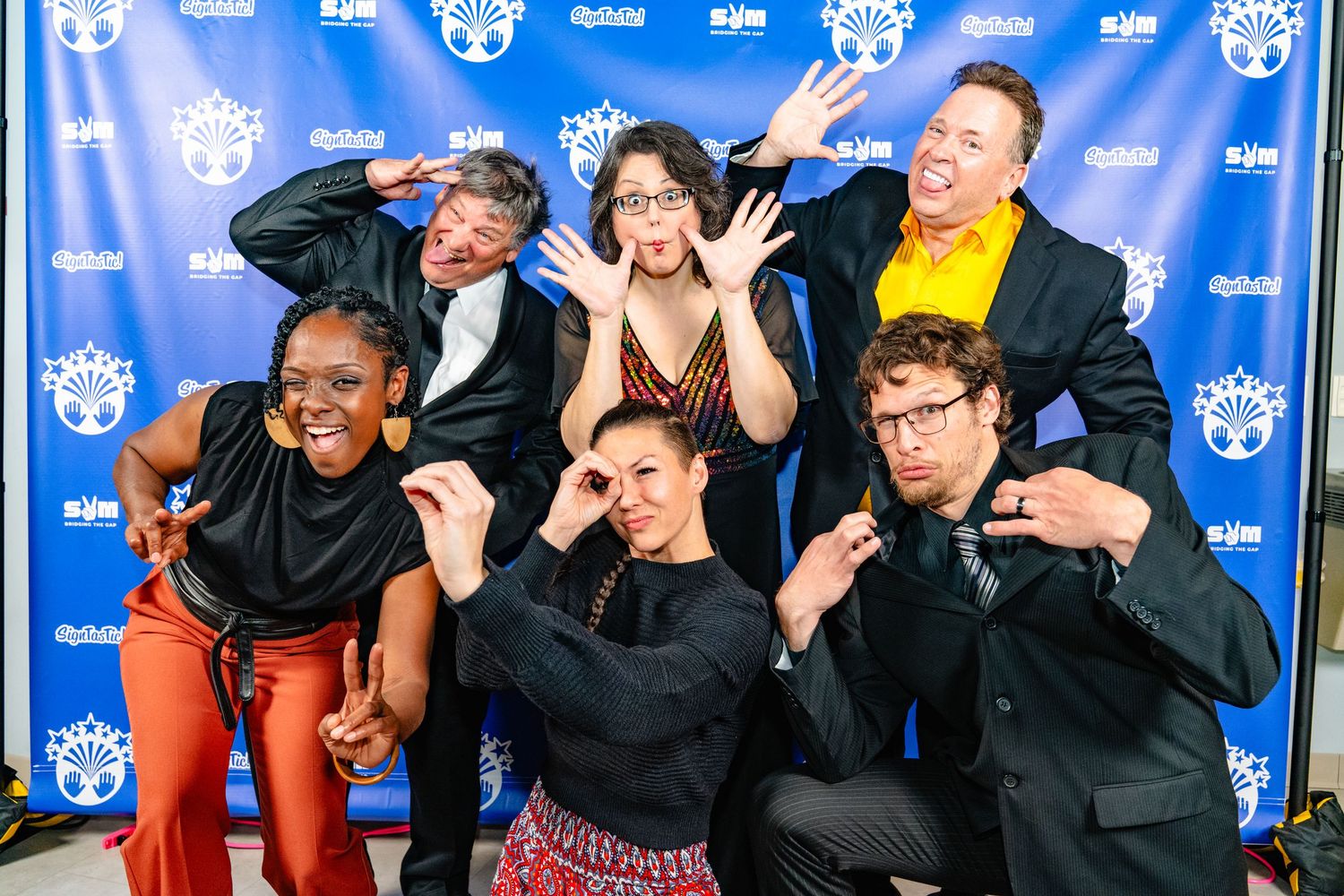 A group of performers pose playfully and make funny faces while posing on a blue backdrop at a press event.