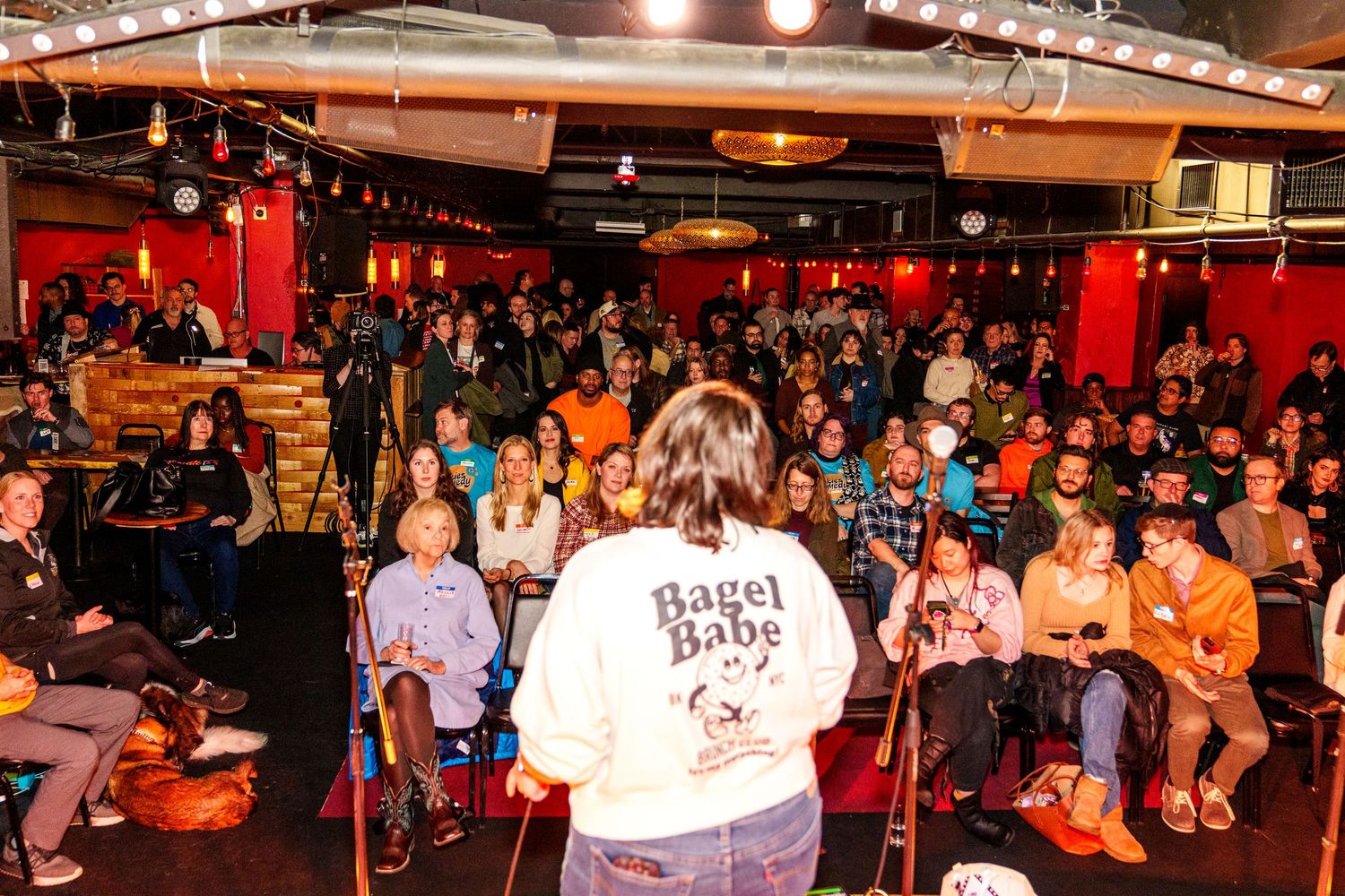 Wide view of a packed indoor event space with string lights and red ambient lighting showing crowd mingling.