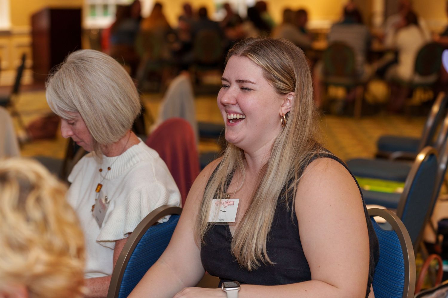 Young woman laughing at a conference event while seated at a table with a name tag.