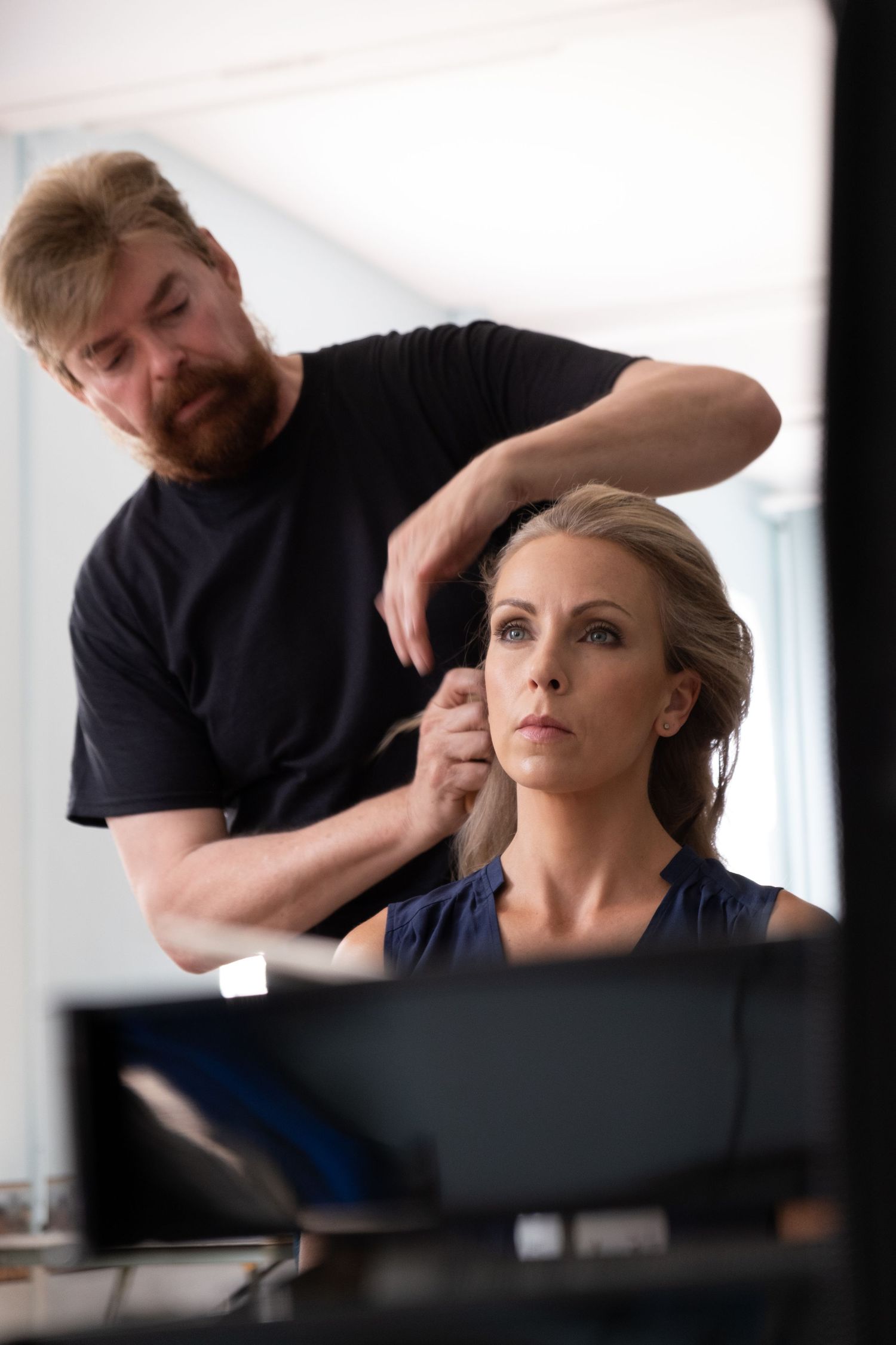 A bearded hairstylist in a black shirt styling a blonde womans hair in a bright studio setting.