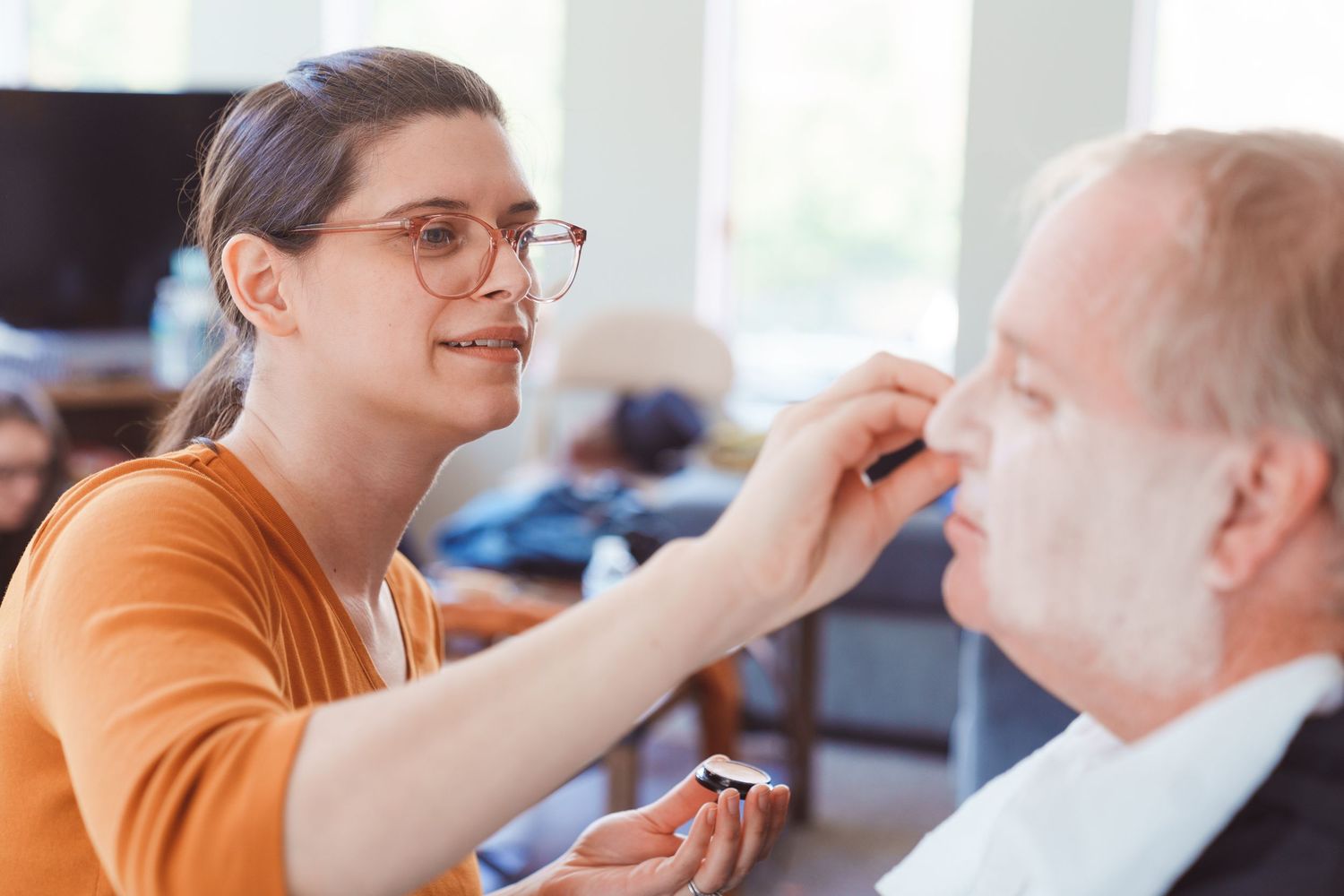 Young woman with glasses applying makeup to an older man in a bright living room setting.