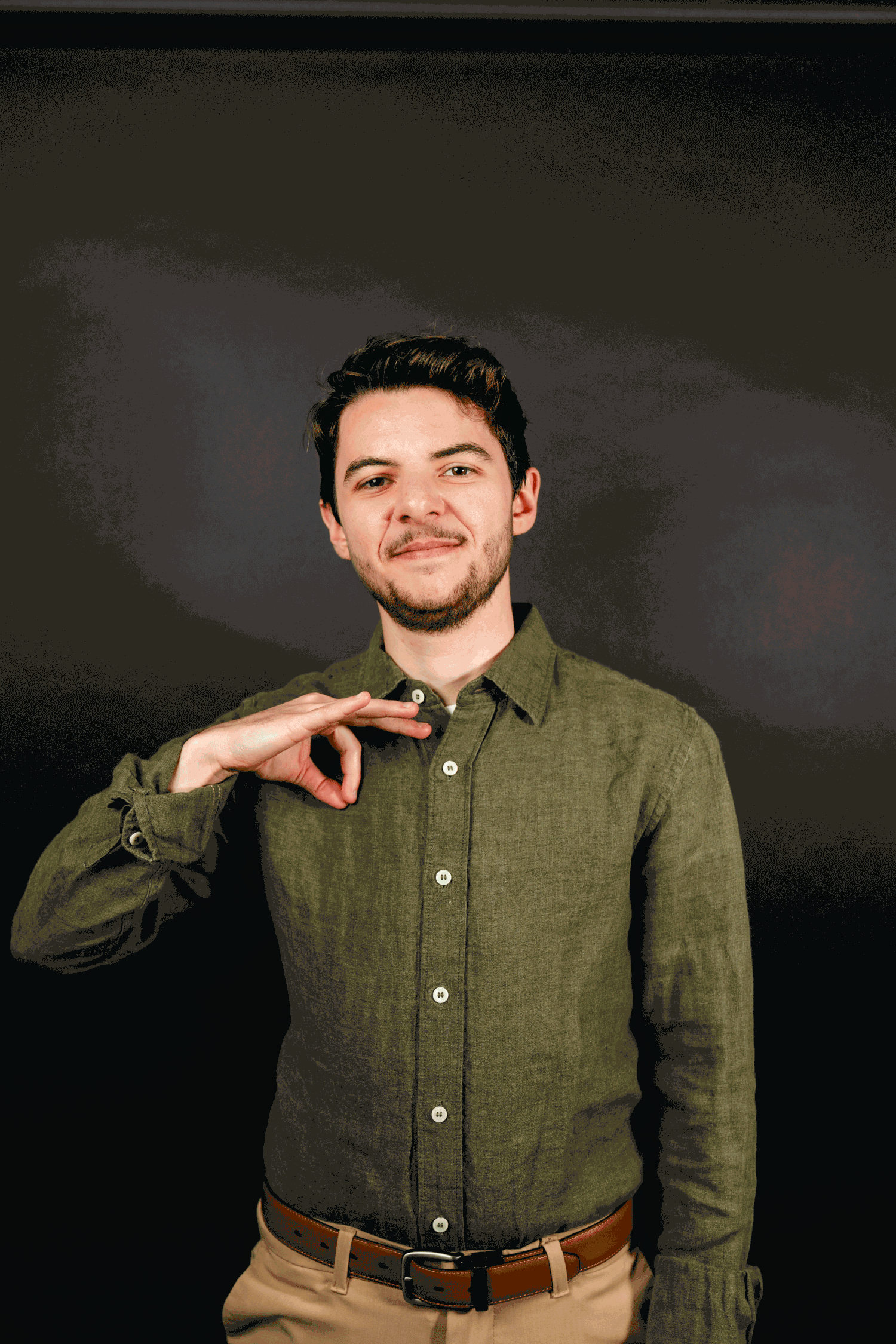 A person in an olive green button-down shirt and khaki pants poses casually against a dark background.