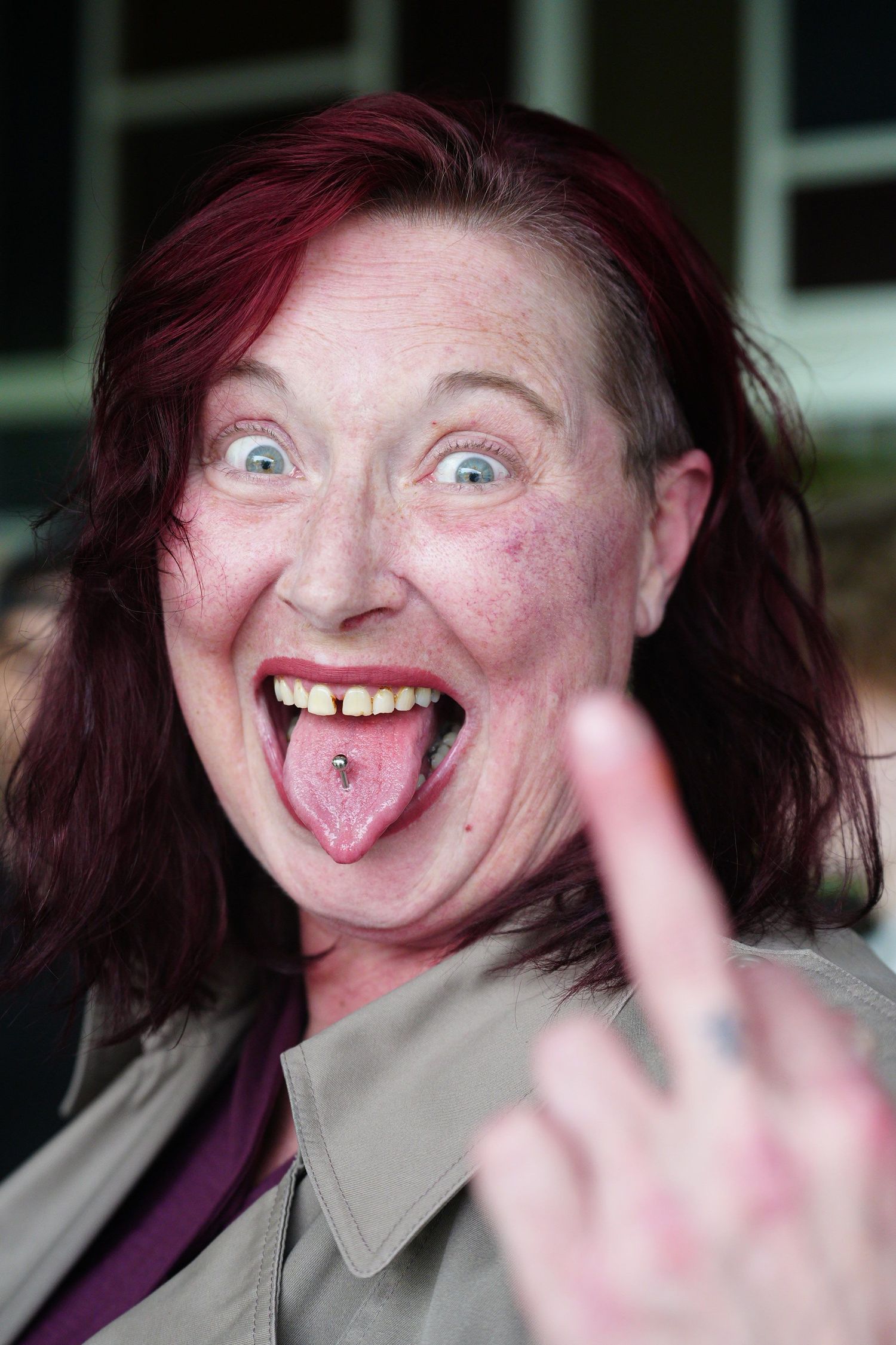 Woman with red hair, tongue piercing, sticking out tongue and giving middle finger gesture outdoors.