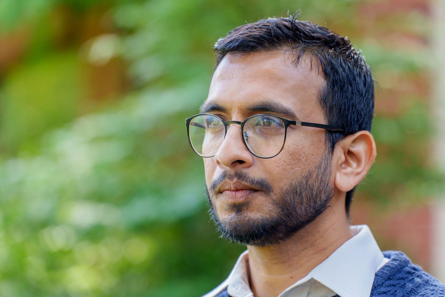 Young man with glasses and beard outdoors, with green foliage background, thoughtful expression.