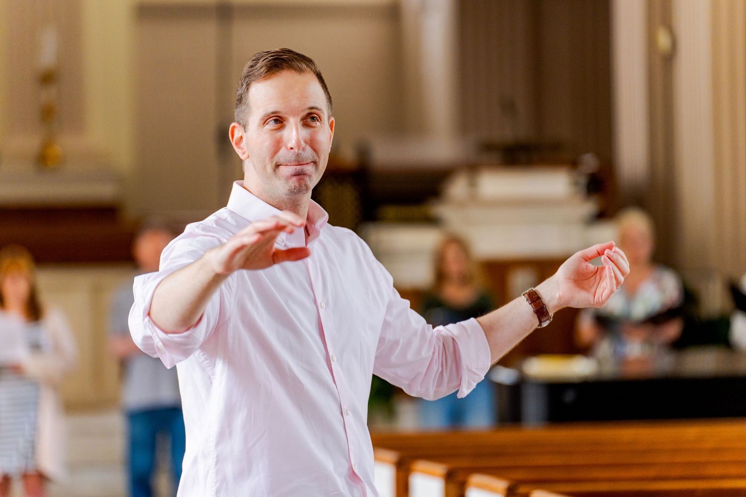 Man in pink shirt conducting or directing in a church hall with people seated in the background.