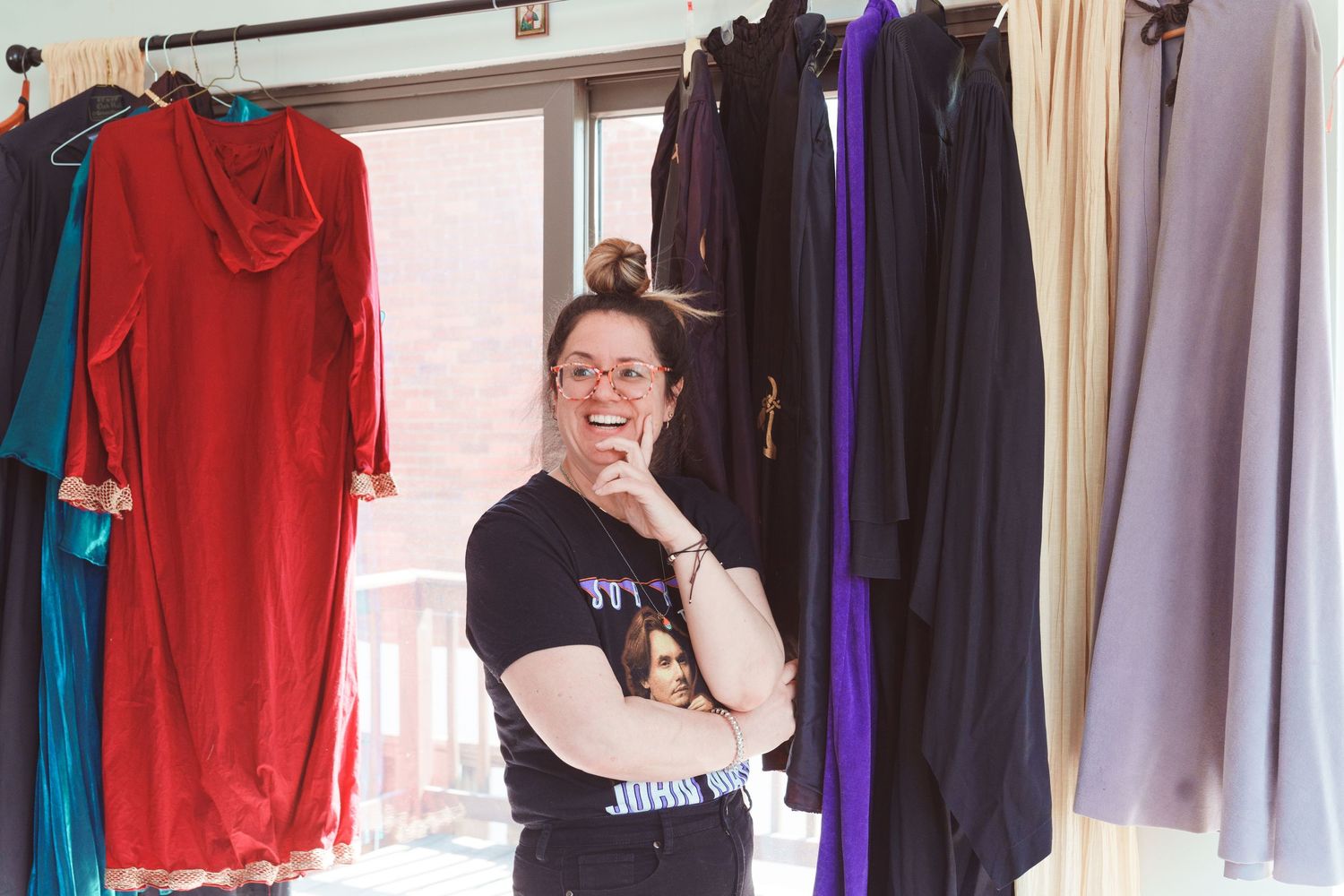 Woman smiling among colorful hanging garments on a clothing rack near a bright window.