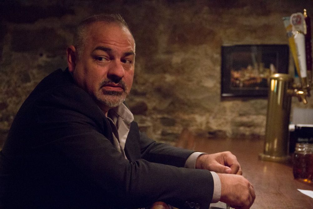 A middle-aged man with a beard sits at a bar in a dimly lit stone-walled room, looking pensively to the side.