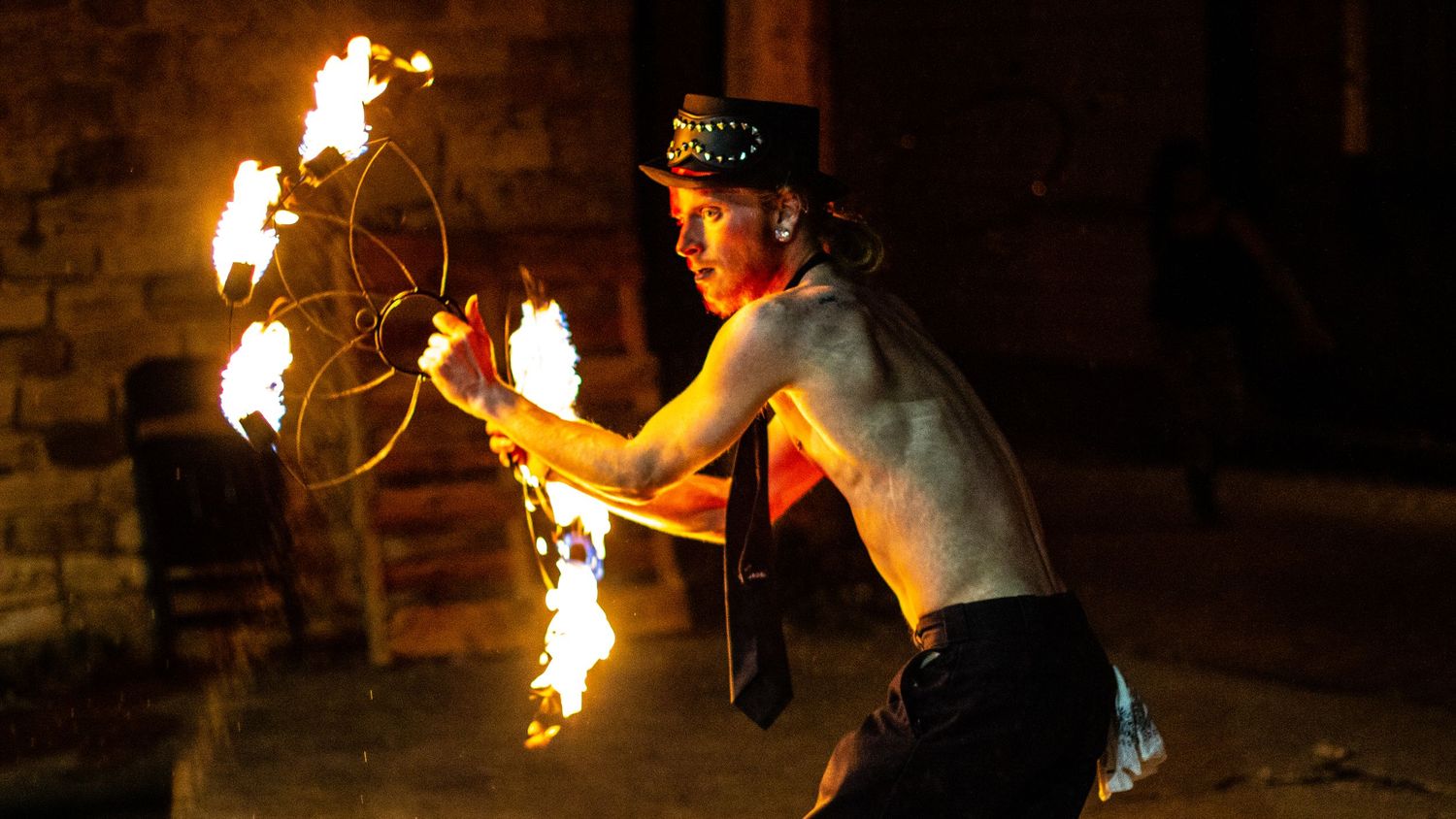 Fire performance artist spinning flames in dark venue with brick wall backdrop.