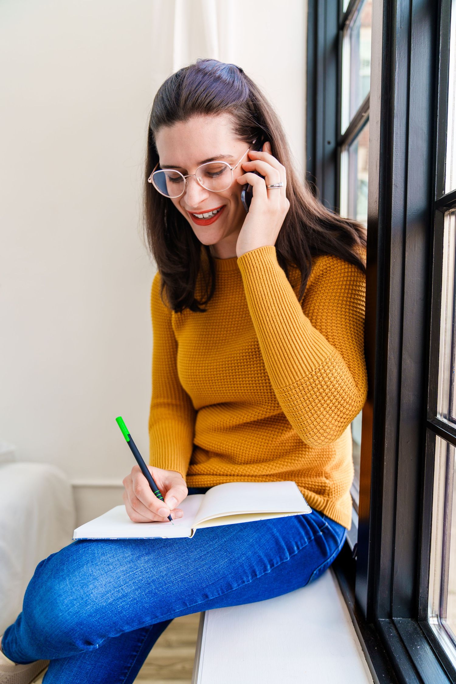 Someone in glasses and a mustard sweater takes notes while sitting by a window.
