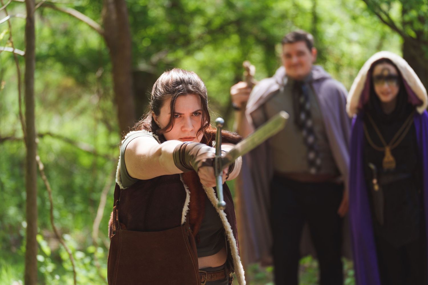 Woman in medieval costume aiming bow and arrow in forest with costumed characters in background.
