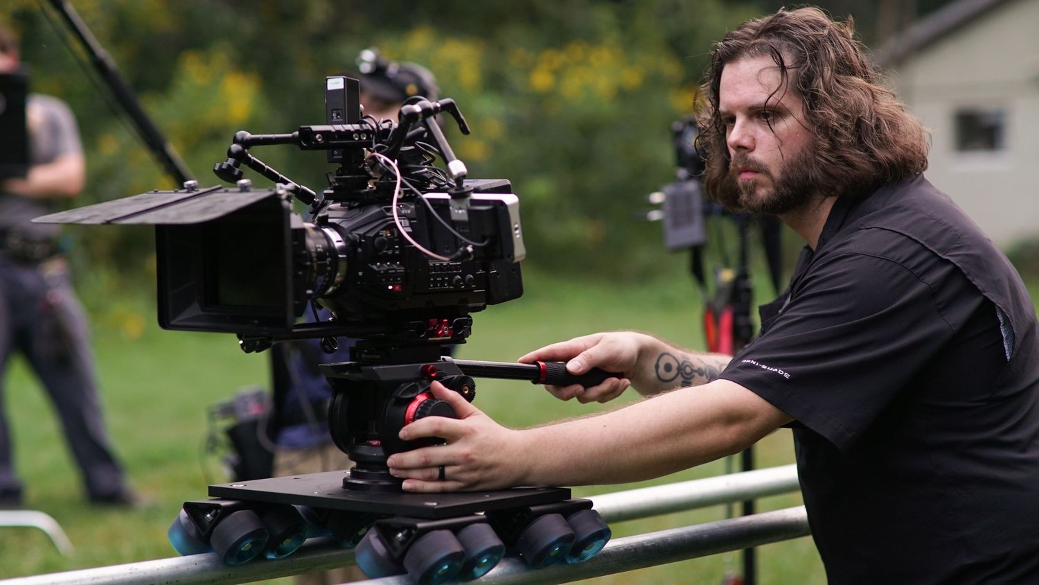 A cameraman with long hair operating a professional film camera on a dolly outdoors.